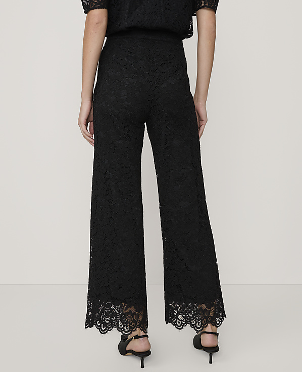 A person stands with their back to the camera wearing black lace wide-leg trousers and heels against a plain, light-colored background.