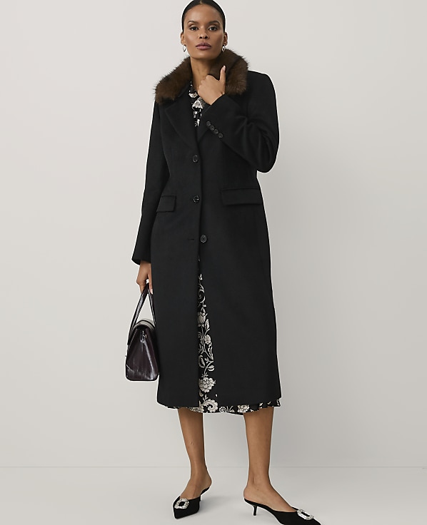 A woman wearing a long black coat with a faux-fur collar and a floral dress stands against a plain white background.