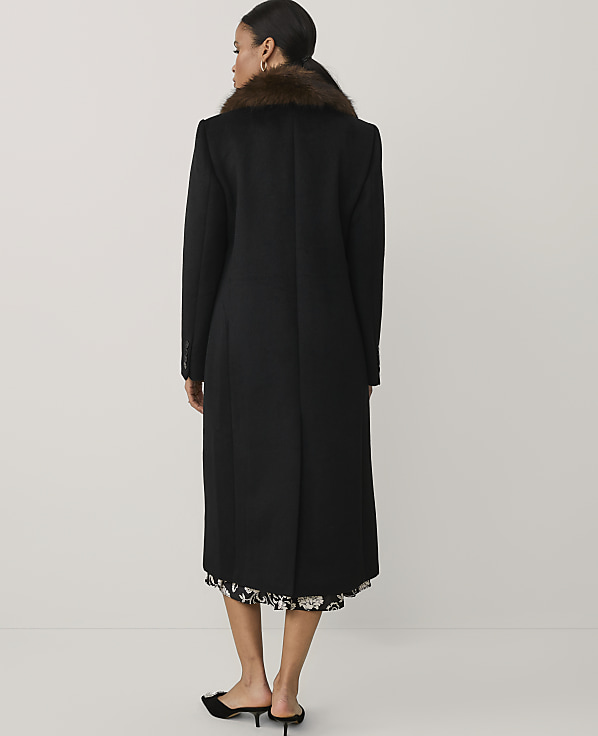 A woman stands with her back to the camera wearing a long black coat with a brown faux-fur collar against a plain, light-colored background.