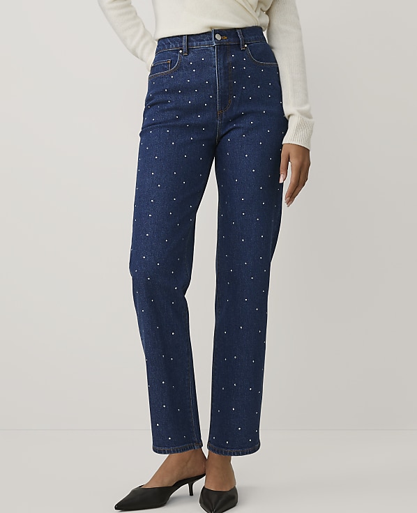 A person wears a white sweater, blue polka-dot jeans, and black heels against a plain light gray background.