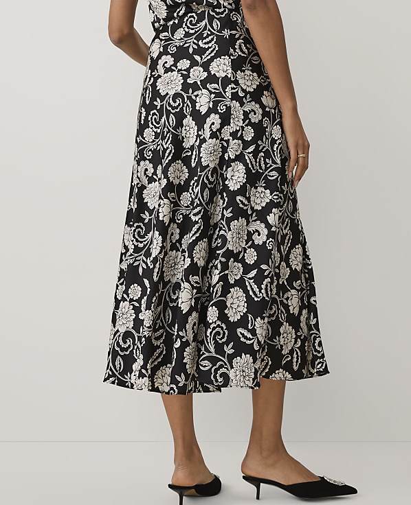 A person wears a black and white floral midi dress and black pointed-toe mules against a plain white background.