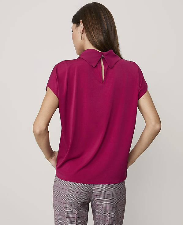 A woman stands with her back to the camera wearing a magenta short-sleeved blouse and plaid trousers against a plain light gray background.