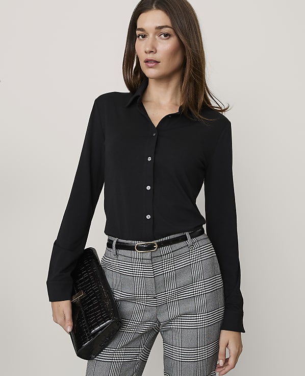 A woman wearing a black button-down shirt and plaid trousers stands against a plain, light-colored background.