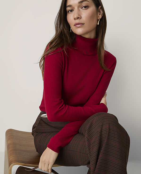 A woman wearing a red turtleneck and plaid trousers sits on a wooden chair against a plain white background.