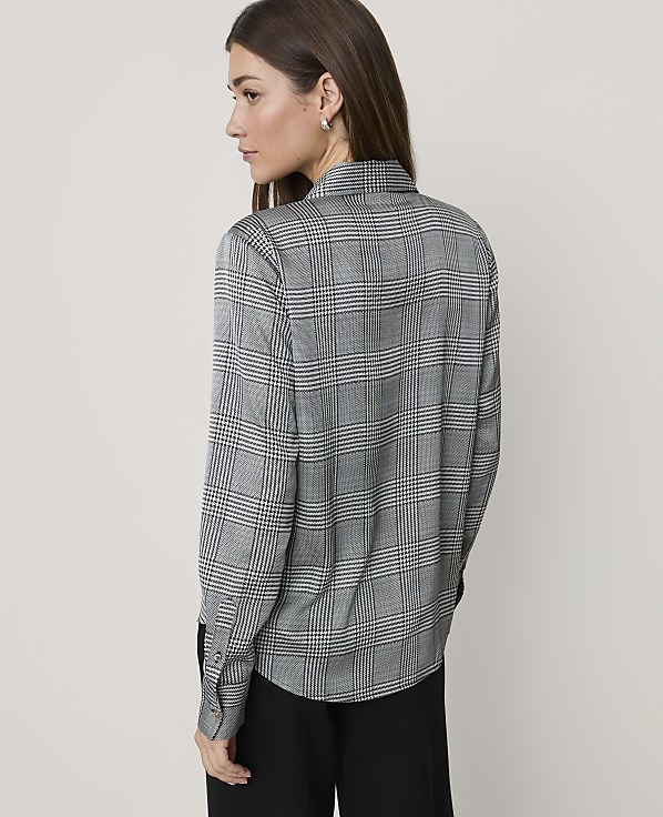 A woman wearing a grey plaid shirt and black trousers stands against a plain, light-colored background.