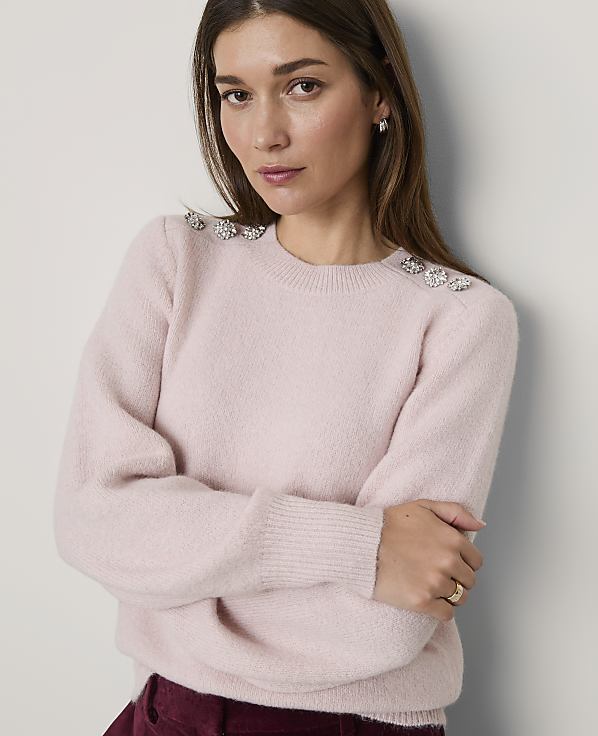 A woman wearing a light pink sweater with jeweled shoulder accents leans against a plain white wall.