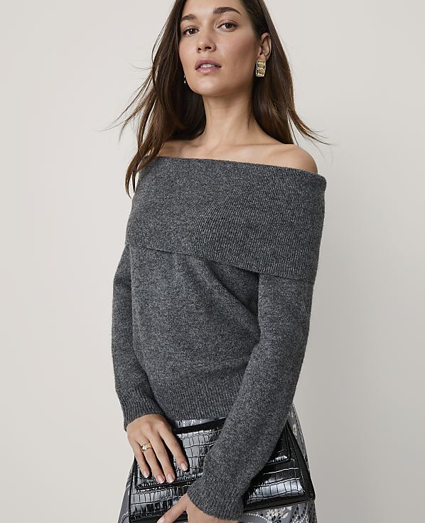 A woman wearing a grey off-the-shoulder sweater and holding a black clutch stands against a plain, light-colored background.