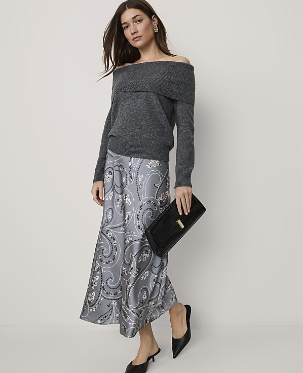 A woman wearing a grey off-the-shoulder sweater and a paisley print skirt stands against a plain, light-colored background.
