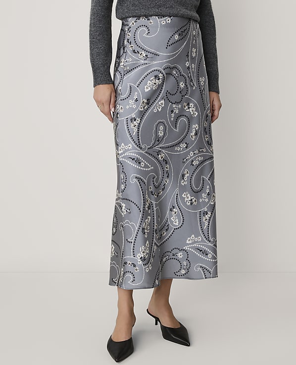 A person wearing a grey sweater, a grey paisley-print midi skirt, and black pointed-toe heels stands against a plain white background.