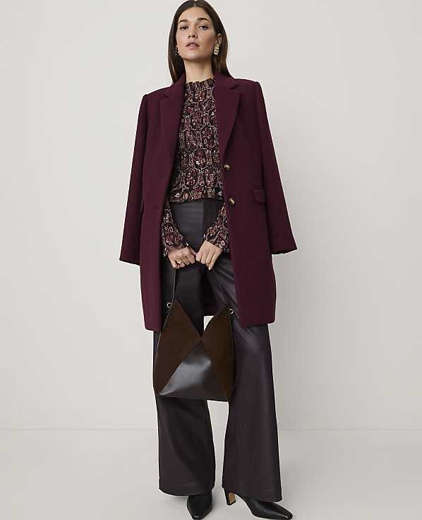 A woman wearing a burgundy coat, patterned blouse, and dark leather trousers stands against a plain white background.