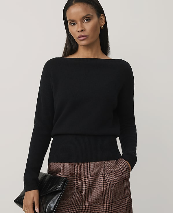 A woman wearing a black sweater and a brown plaid skirt holds a black clutch against a plain light gray background.