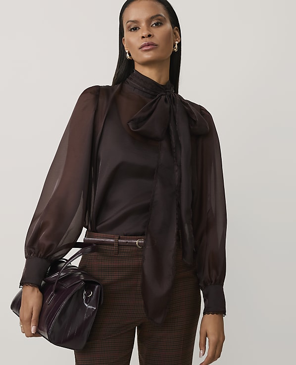 A woman wearing a sheer brown pussy-bow blouse and checkered trousers stands against a plain light grey background.