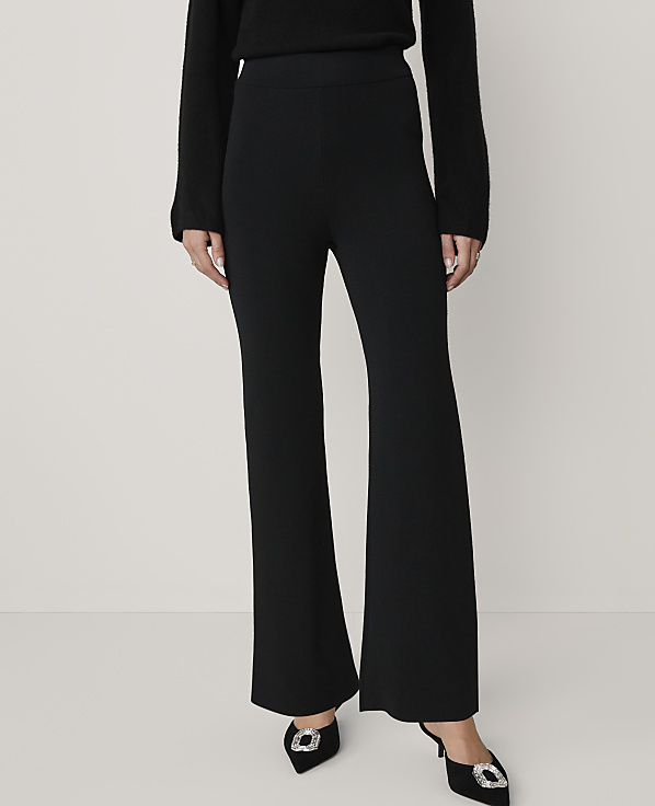 A person wearing a black sweater, black wide-leg trousers, and black pointed heels with buckles stands against a plain light gray background.