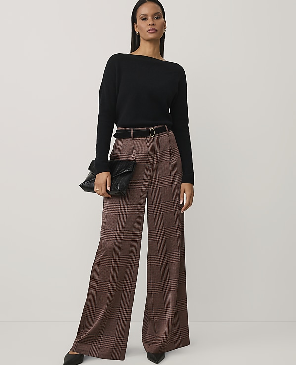 A woman wearing a black sweater and brown plaid wide-leg trousers stands against a plain, light-colored background.