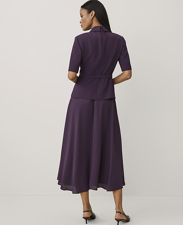 A woman stands with her back to the camera wearing a purple short-sleeved jacket and matching long skirt against a plain white background.