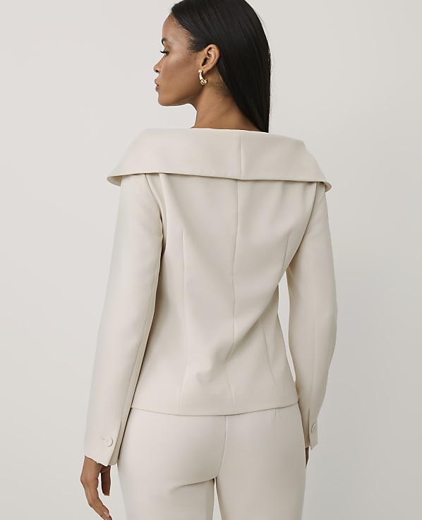 A woman wearing a cream-colored blazer and matching trousers stands with her back to the viewer against a plain, light-colored background.