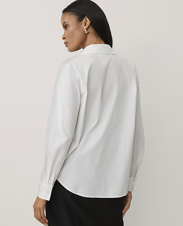 A woman wearing a white long-sleeved button-down shirt and black trousers stands against a plain, light-colored background.