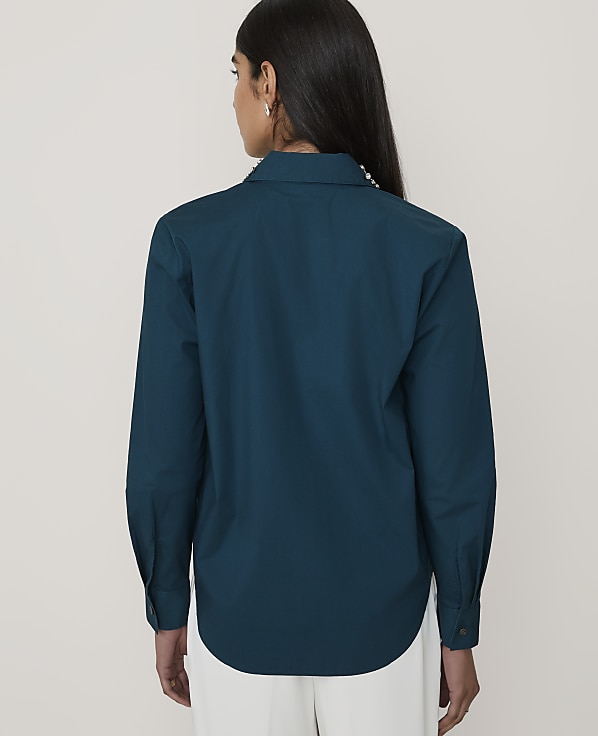 A woman wearing a teal long-sleeved shirt and white trousers stands with her back to the camera against a plain, light-colored background.
