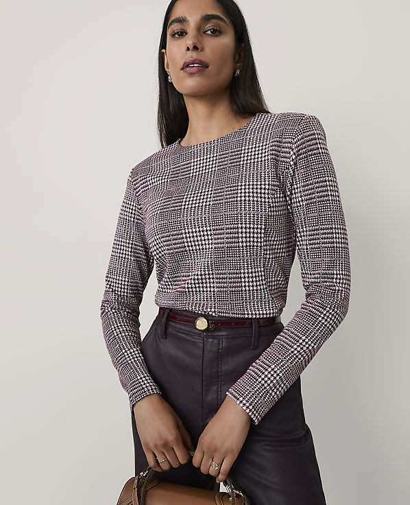 A woman wearing a maroon and white houndstooth top and dark leather trousers stands against a plain, light-colored background.
