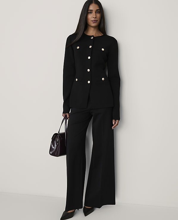 A woman wearing a black buttoned blazer and wide-leg trousers holds a burgundy handbag against a plain white background.