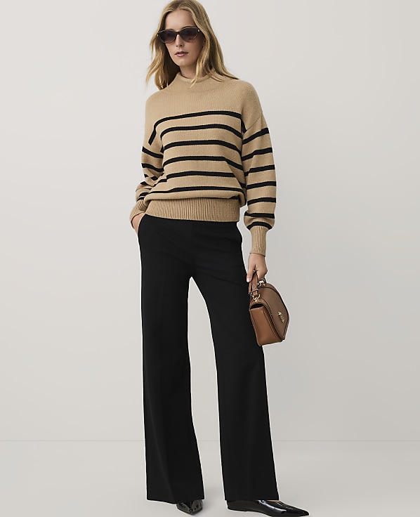 A woman wearing a tan striped sweater, black wide-leg trousers, and sunglasses stands against a plain white background.