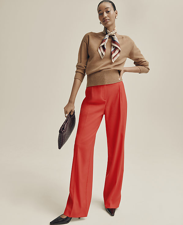 A woman wearing a tan sweater, a patterned neck scarf, and wide-leg red trousers stands against a plain, neutral-toned background.