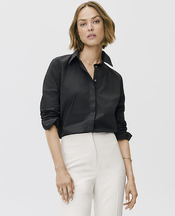 A woman wearing a black button-down shirt and white trousers stands against a plain light gray background.