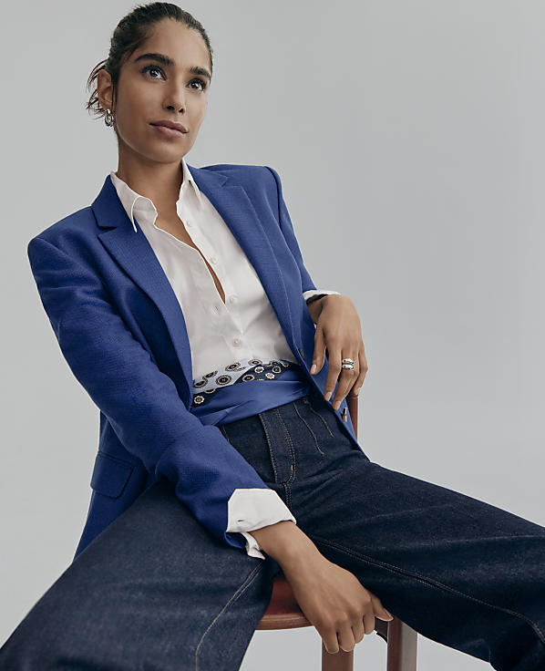 A woman wearing a blue blazer, white shirt, and dark jeans sits on a wooden stool against a plain light gray background.
