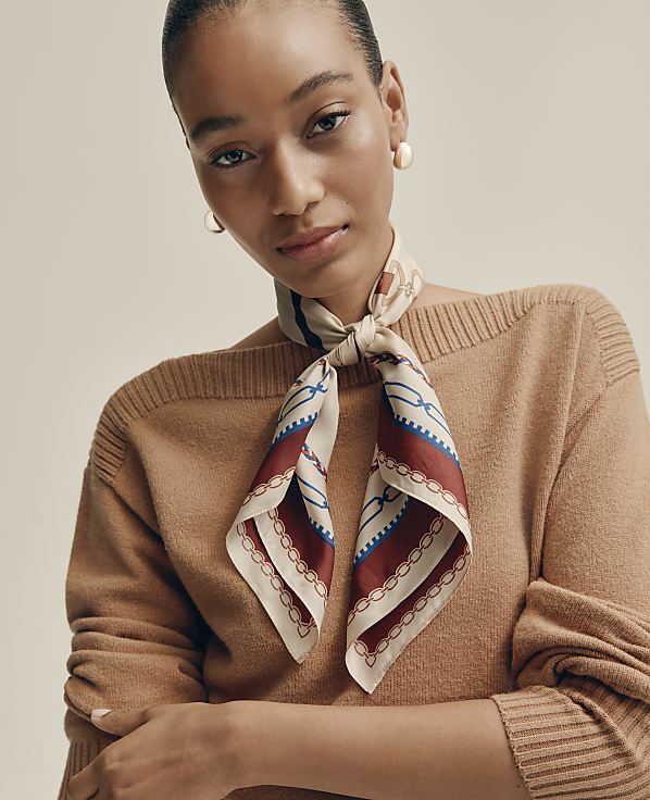 A woman wearing a brown sweater and a patterned silk scarf poses against a plain, neutral-toned background.