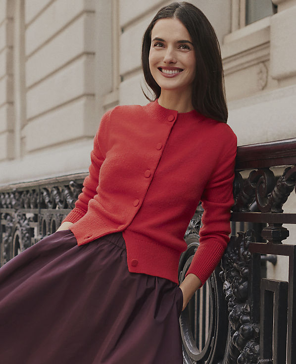 A smiling woman wearing a red cardigan and a dark purple skirt leans against an ornate black iron railing in front of a light-colored stone building.