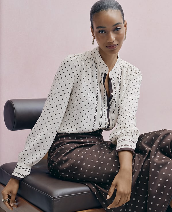 A woman wearing a patterned white blouse and brown patterned trousers sits on a dark leather bench against a plain pink background.