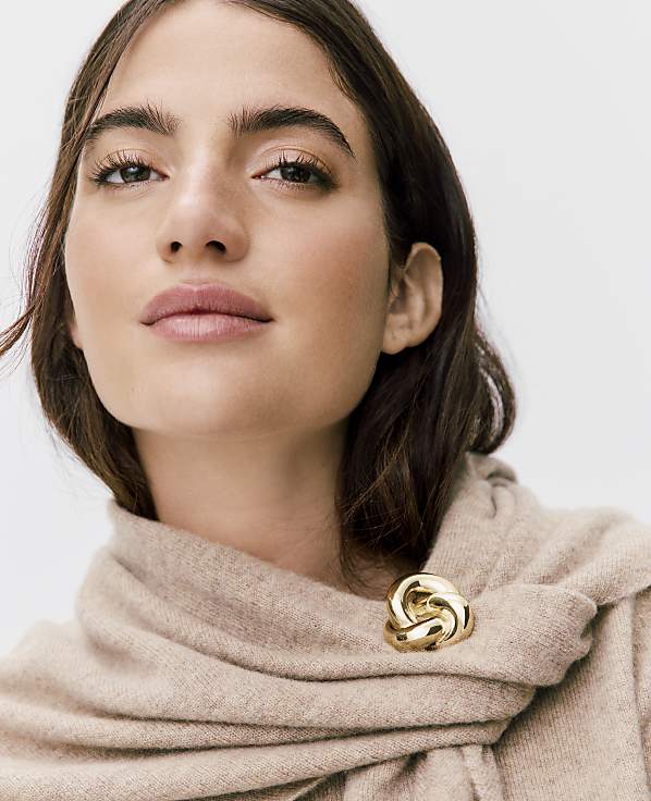 A woman with brown hair and a beige scarf fastened with a gold brooch poses against a plain white background.