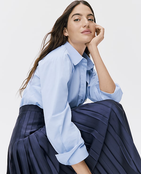 A woman wearing a light blue button-down shirt and a dark navy pleated skirt poses against a plain white background.