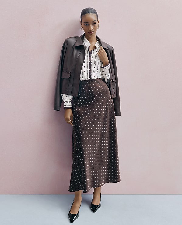 A woman wearing a brown leather jacket, a patterned white shirt, and a polka-dot brown skirt stands against a plain pink wall.