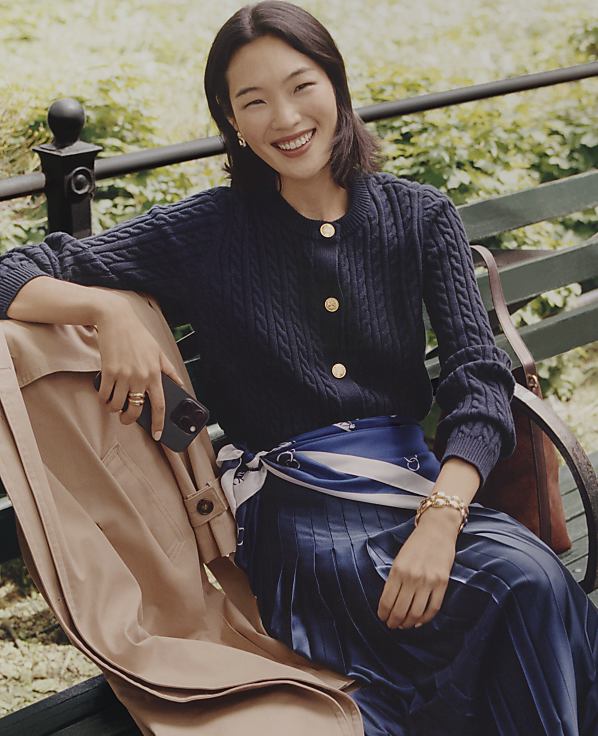 A smiling woman in a navy cable-knit cardigan and pleated skirt sits on a green park bench with a tan coat beside her, set against a backdrop of lush greenery.