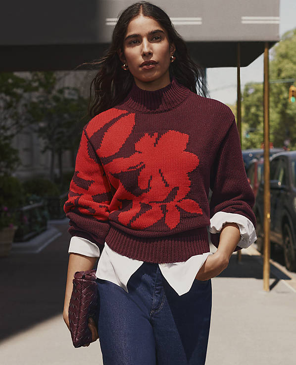 A woman wearing a maroon floral sweater and blue jeans stands on a city sidewalk with cars and greenery in the background.