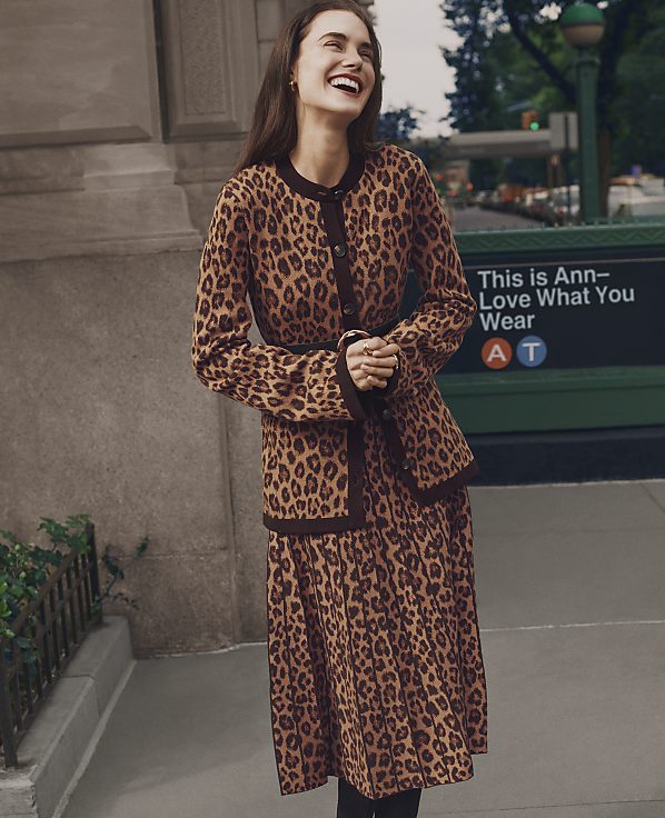 A laughing woman wearing a leopard-print outfit stands on a city sidewalk in front of a stone building and a green subway entrance sign.