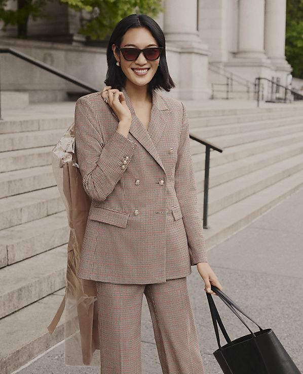 A smiling woman in a brown checkered suit stands on stone steps in front of a building with large white columns.