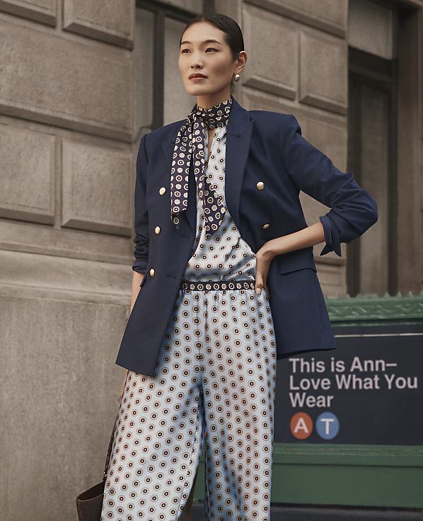 A woman wearing a patterned blue and white outfit with a navy blazer stands before a stone building and a green sign.