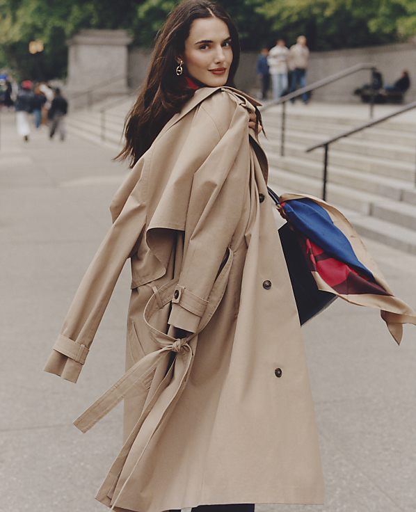 A woman in a tan trench coat stands in the foreground against a blurred city background with stone steps and pedestrians.