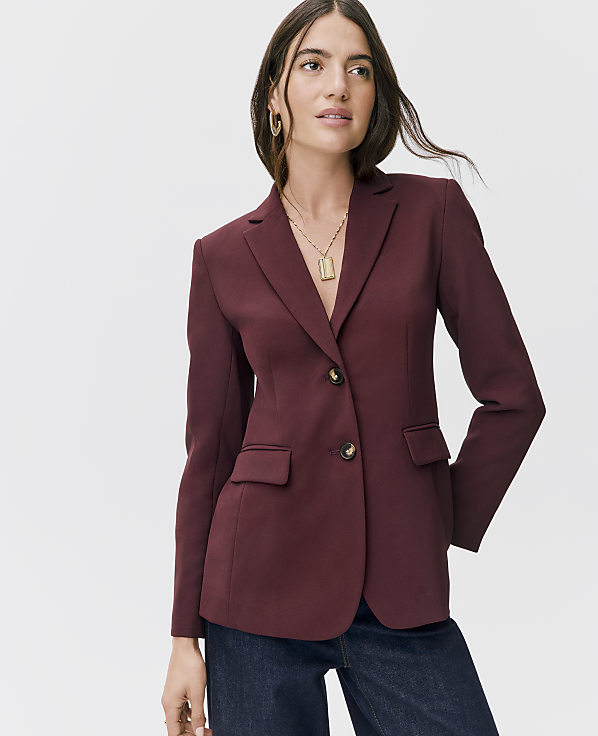 A woman wearing a maroon blazer and dark blue jeans stands against a plain white background.