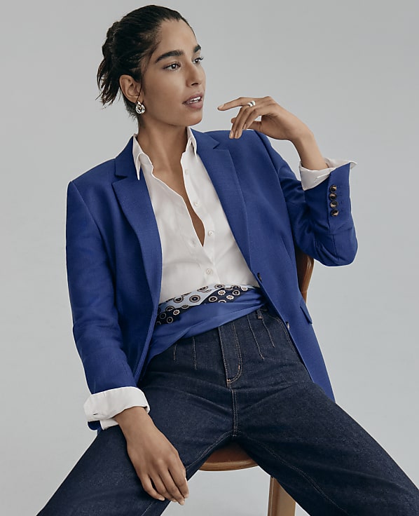 A woman wearing a blue blazer, white shirt, and dark jeans sits on a wooden chair against a plain grey background.