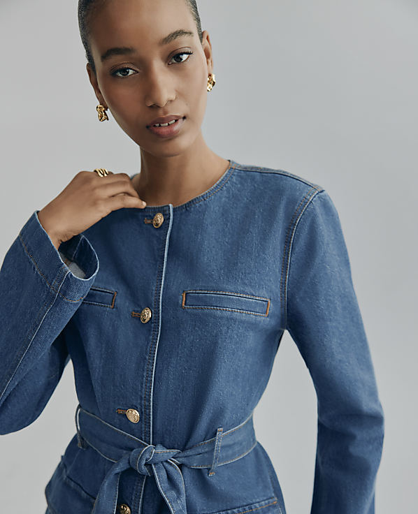 A woman wearing a blue denim jacket and gold earrings poses against a plain grey background.