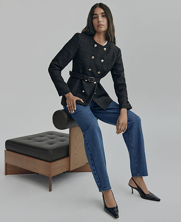 A woman wearing a black double-breasted blazer and blue jeans sits on a modern black leather and wood bench against a plain grey background.