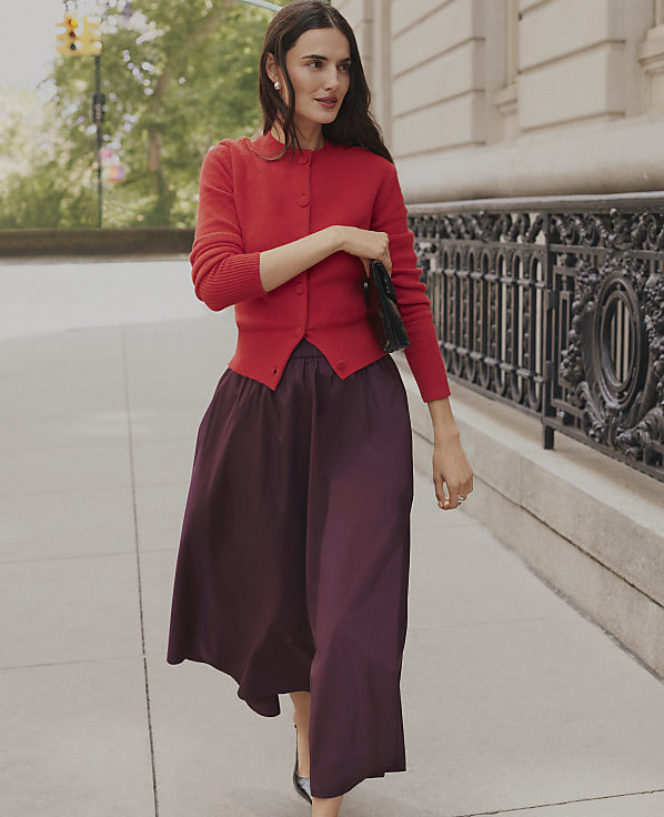 A woman wearing a red cardigan and a long purple skirt walks along a city sidewalk next to an ornate black iron fence.