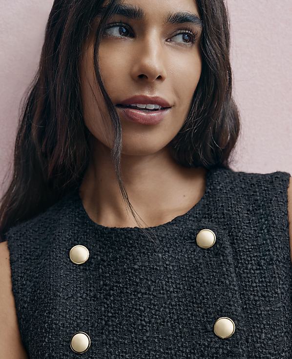 A woman with long dark hair wearing a black textured jacket with gold buttons poses against a light pink wall.