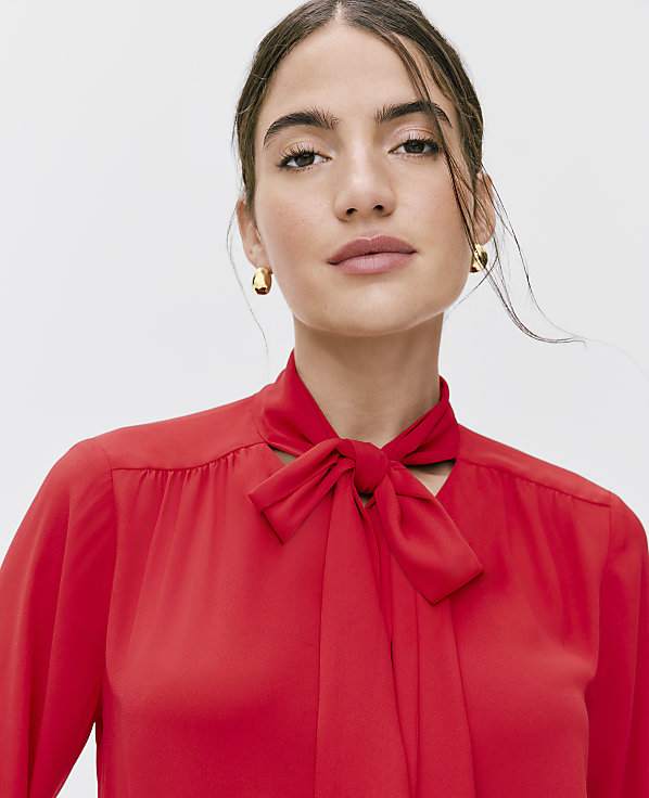A woman wearing a red bow-tie blouse and gold earrings poses against a plain white background.
