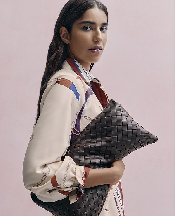 A woman in a patterned cream blouse holds a large woven brown leather clutch against a solid pink background.