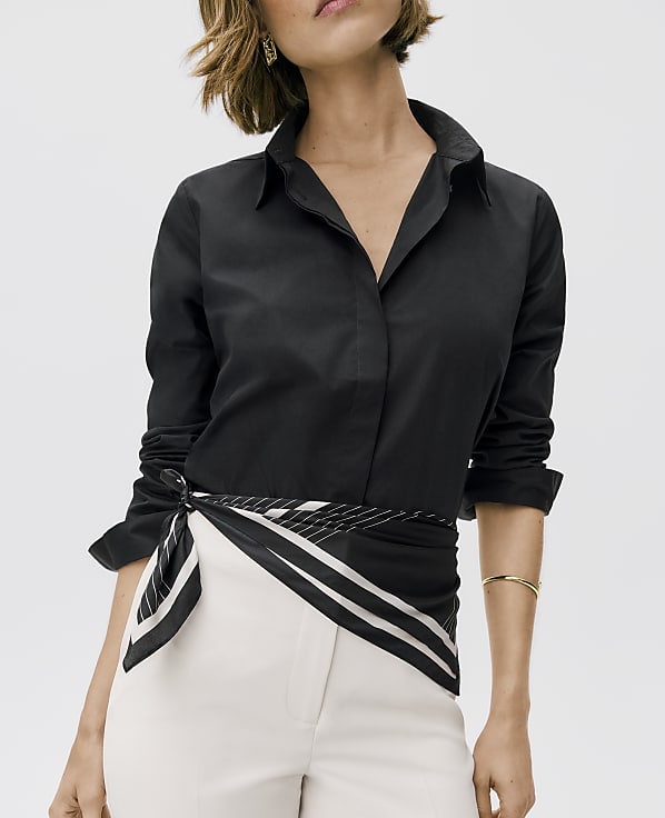 A woman wears a black button-down shirt and white trousers with a patterned scarf tied at the waist against a plain white background.
