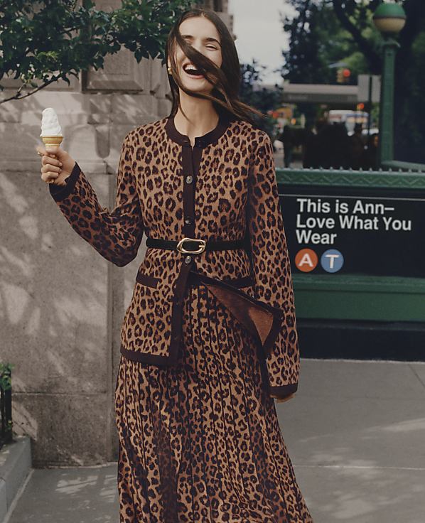 A laughing woman in a leopard-print dress holds an ice cream cone on a city sidewalk with a green sign and trees in the background.